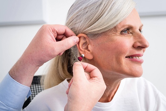 Ältere Frau bekommt Hörgerät angepasst, Hörakustiker unterstützt beim Einsetzen des Geräts.